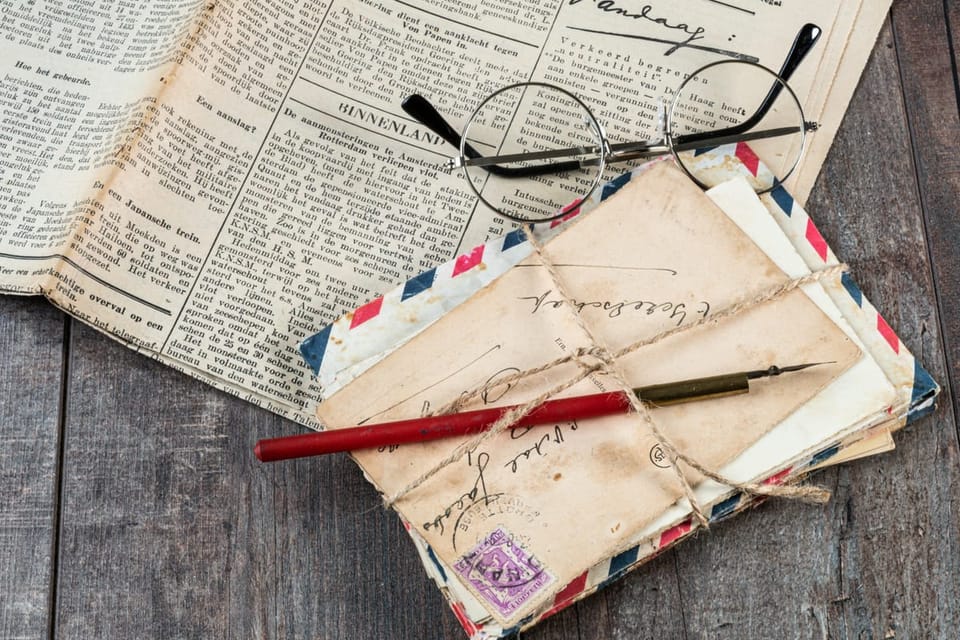 A pile of old fashioned letters, tied up with string with a pen on top. A newspaper and glasses in the background